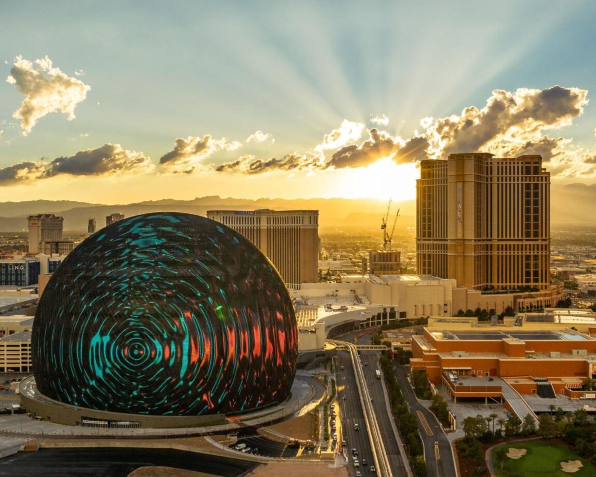 Hotel The Palazzo at The Venetian® Las Vegas Esterno foto The image shows a striking view of a large, spherical building illuminated with colorful patterns. It is set against a sunset backdrop, with rays of sunlight breaking through the clouds. The surrounding cityscape features tall buildings, some under c
